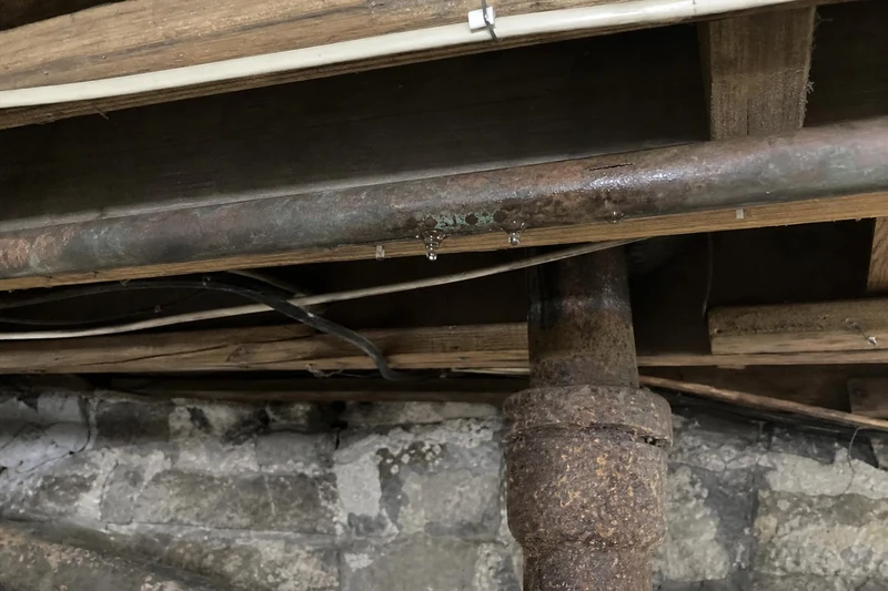 Corroded braided supply line beside a rusted cast-iron stack in the crawlspace of an older Salt Lake City house, rubble stone foundation behind it