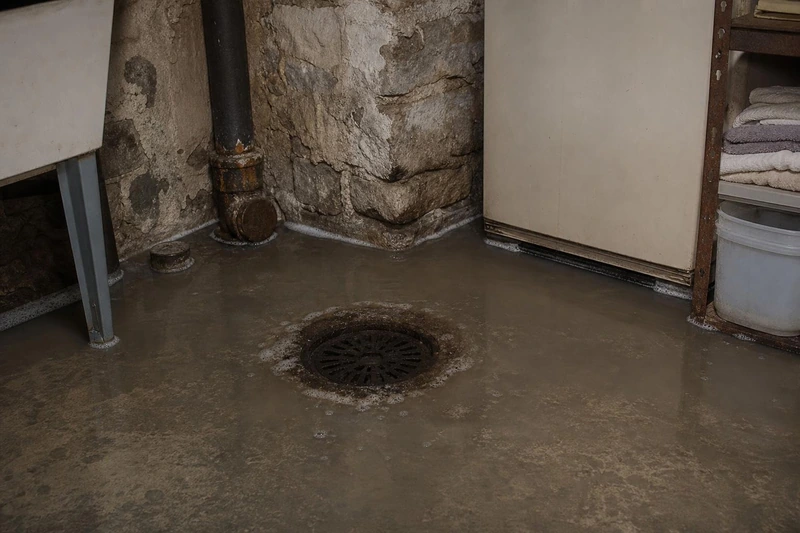 Gray water spreads around a cast-iron floor drain in a Bountiful basement corner, silt ringing it and old stone walls behind