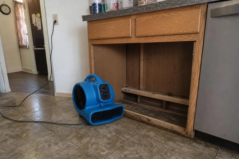 An opened base cabinet sits with its doors off in a Midvale kitchen while an air mover blows into the damp framing
