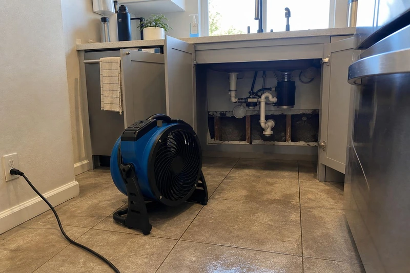 A cut-out cabinet opens on wet framing and a P-trap as a blue mover on the tile blows air inside